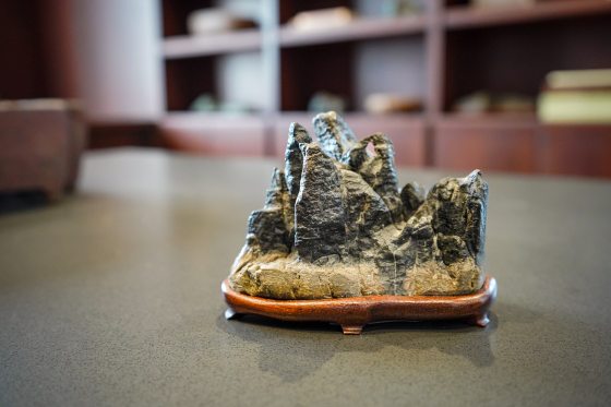  The alt text for this image should be:  A close-up, low-angle shot of a small, dark, craggy rock formation carving, resembling miniature mountains or peaks. It sits on a smooth, dark wooden stand with curved feet. The object is centered on a grey, textured table, with a blurred background of dark wooden shelving or cabinetry.